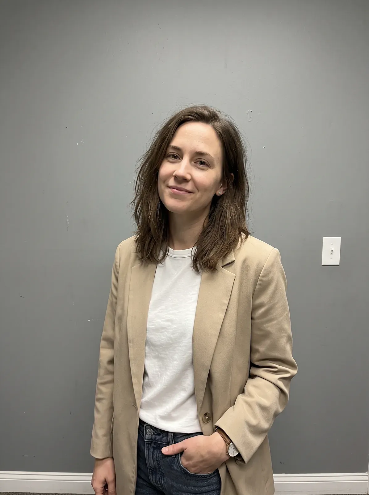 Женщина в бежевом пиджаке и белой футболке стоит на фоне серой стены с выключателем